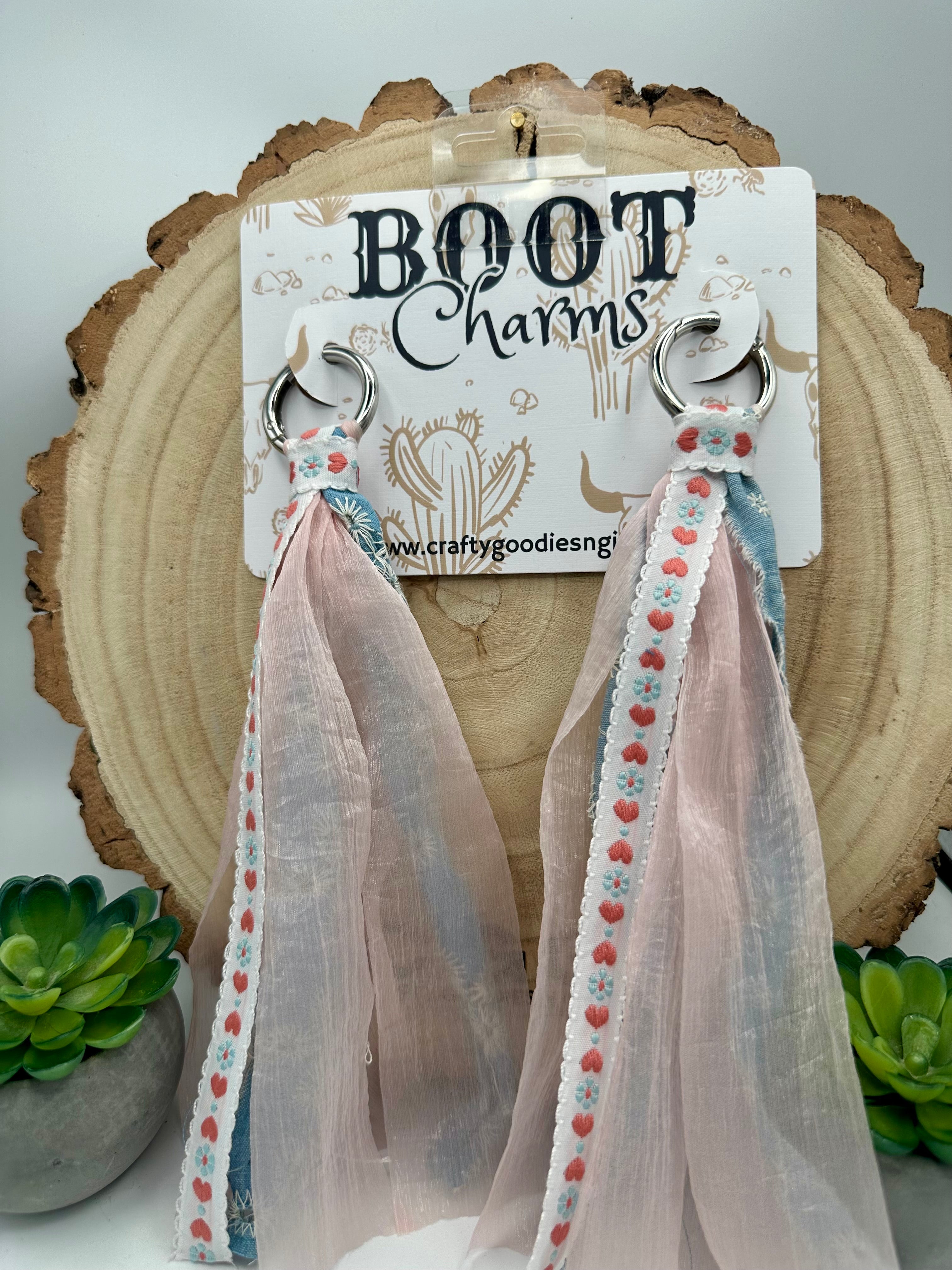 A pair of handmade boot charms that  have silver-tone rings, blush pink sheer ribbon tassels, and white trim with small pink and blue floral details.
