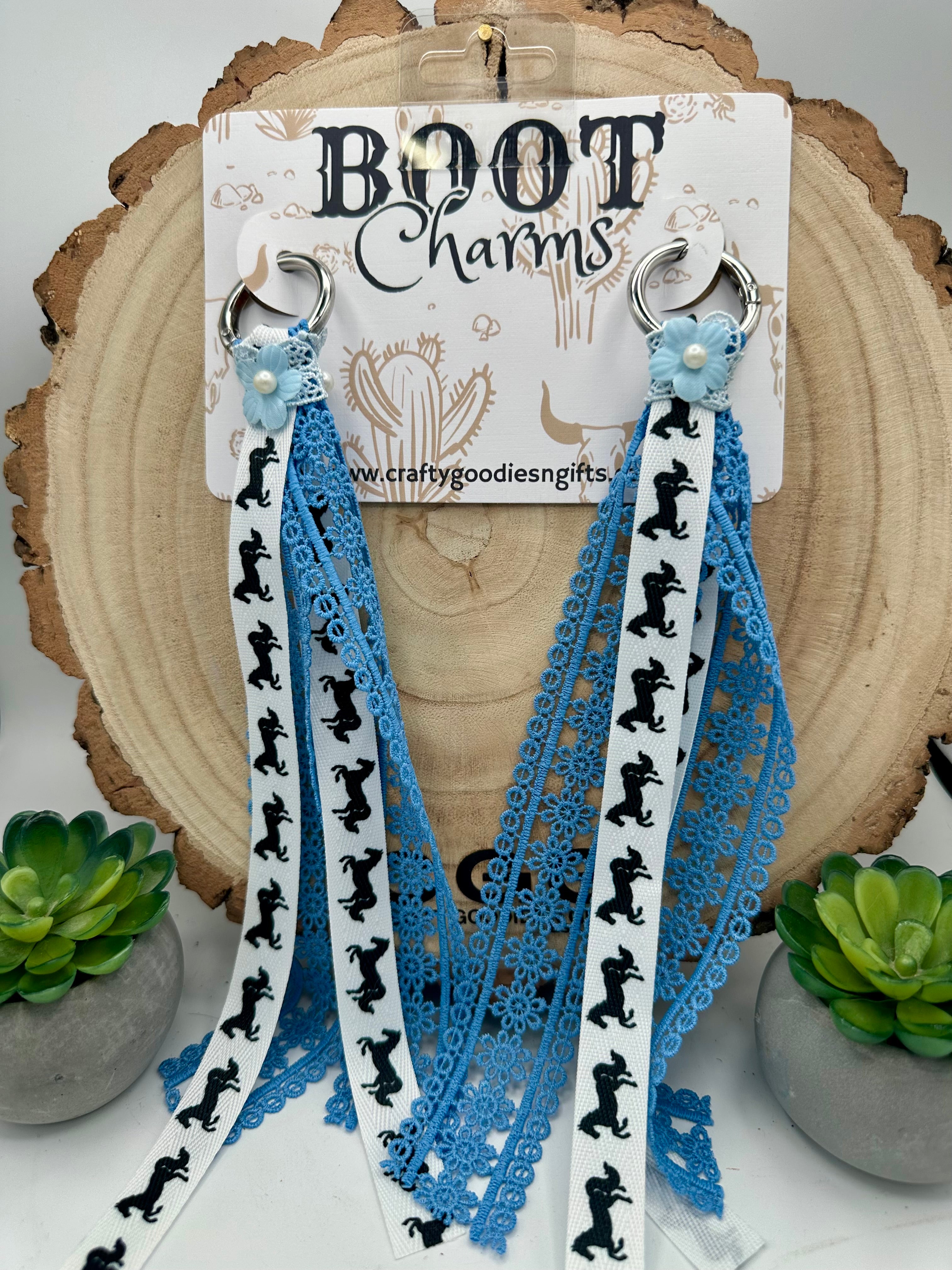 A pair of boot charms made from white and light blue ribbons with black boot silhouettes, blue lace, and small pearl-like accents.