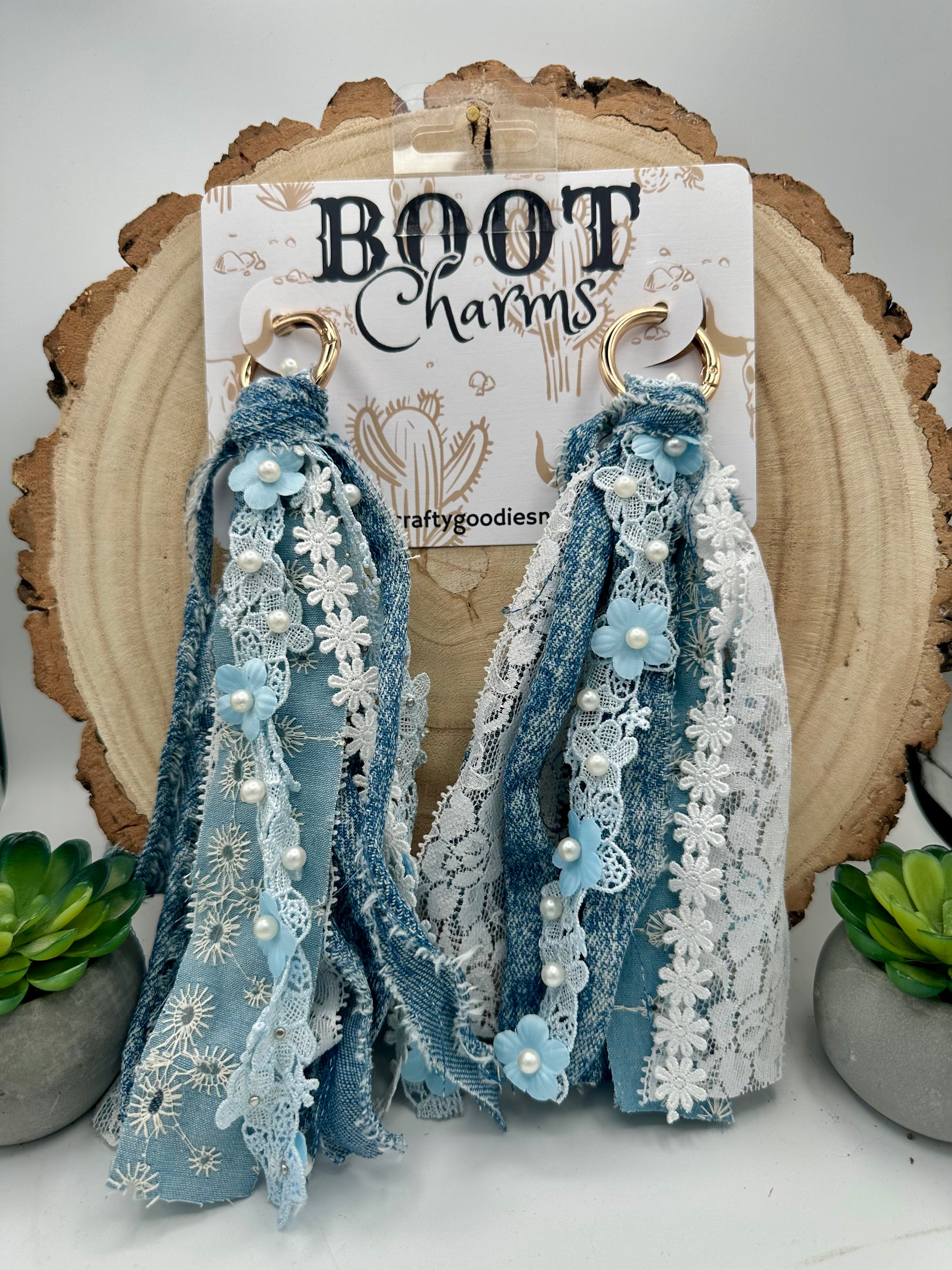 A pair of handmade boot charms featuring layered blue and white lace, denim-like fabric strips, white floral lace, faux pearls, and light blue fabric flowers, hanging from gold-tone rings.
