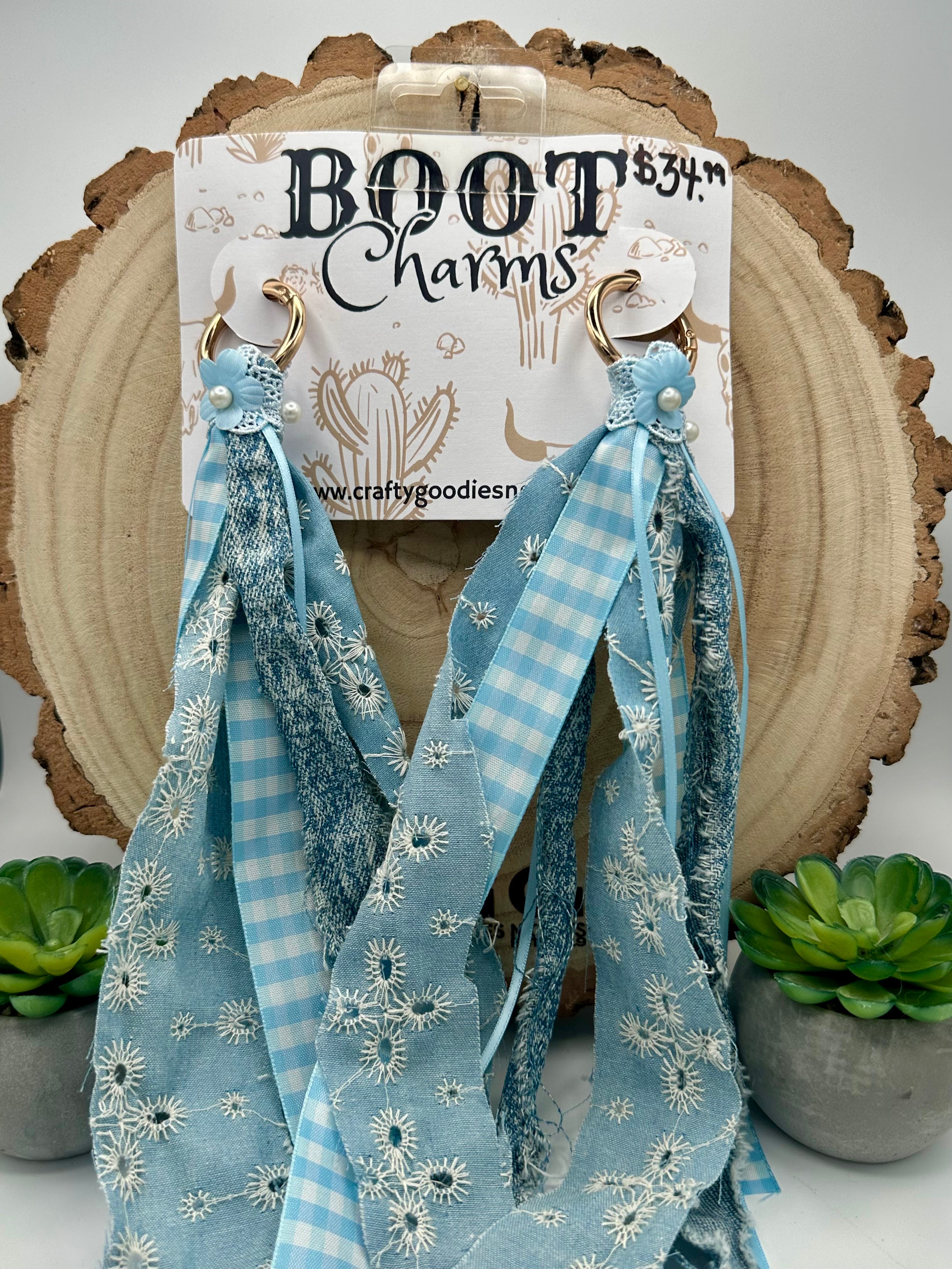 A pair of blue ribbon boot charms with white floral and gingham patterns, hanging from gold rings on a branded cardboard display card.