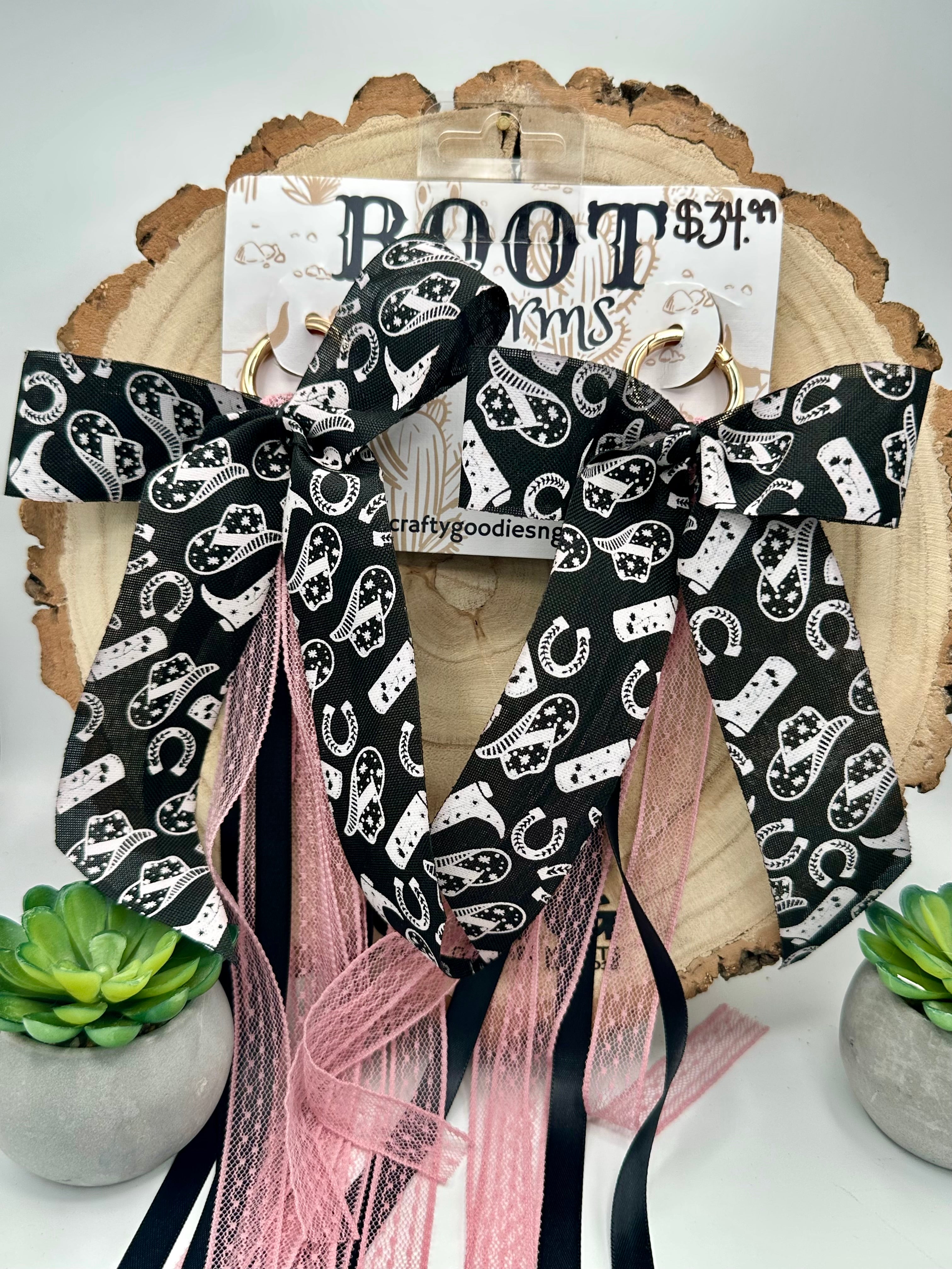 This Set of bot charms have a  large bow with a black and white paisley pattern, featuring pink sheer mesh ribbons and black ribbon tails, displayed on a rustic wood slice with a price tag and small succulent plants.