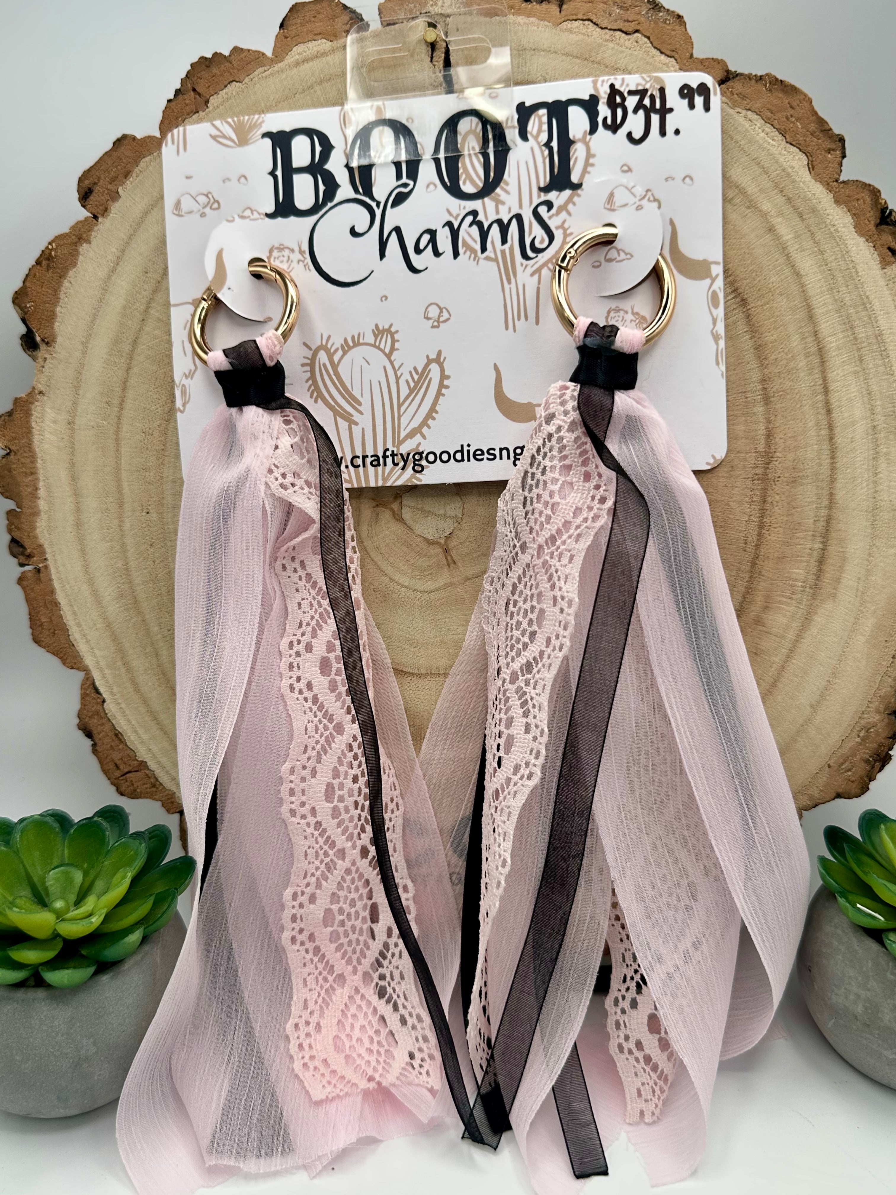 A pair of long boot charms with pale pink tulle, white lace, and dark brown ribbon straps, hanging from gold rings.