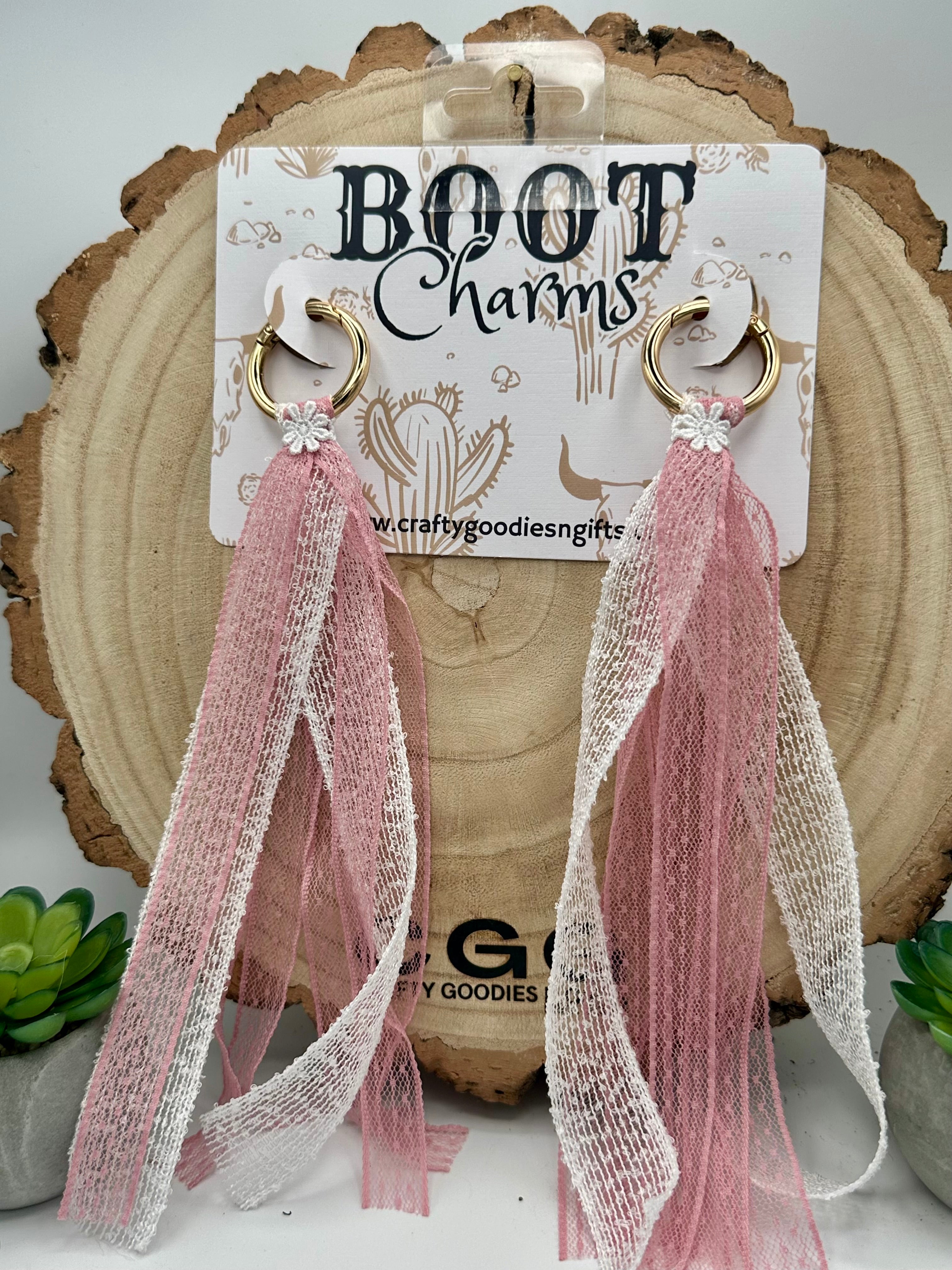 A pair of boot charms with gold-tone hoop tops, small white floral embellishments, and long sheer ribbon tassels in blush pink and white.